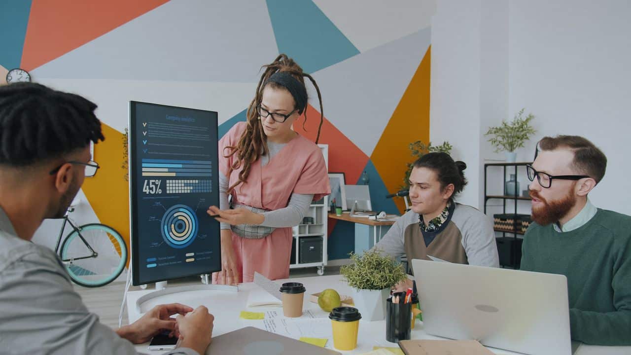 About A diverse team discusses analytics displayed on a monitor in a vibrant office setting, promoting collaboration.